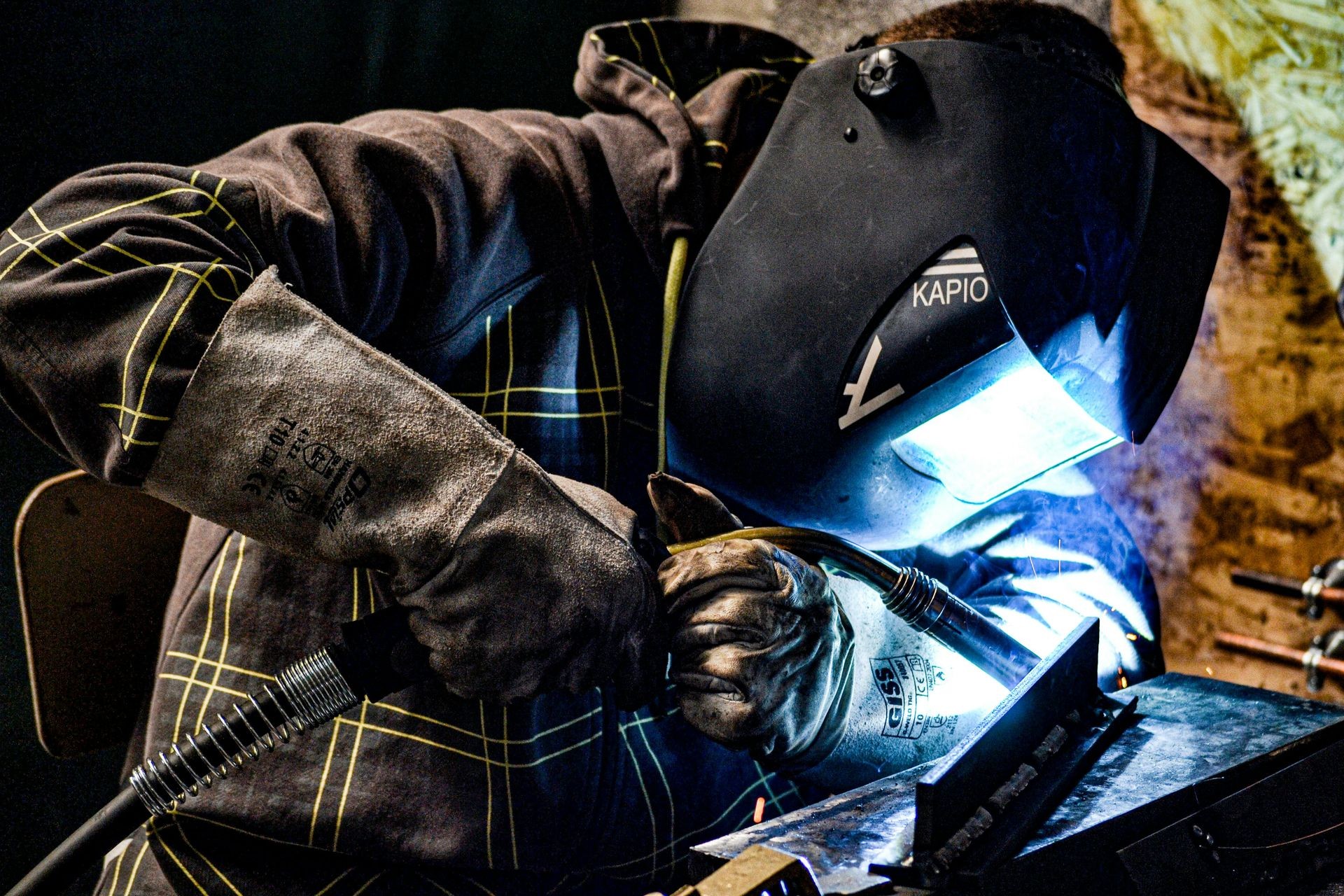 Welder in protective gear working with welding torch emitting bright light and sparks onto a metal surface.