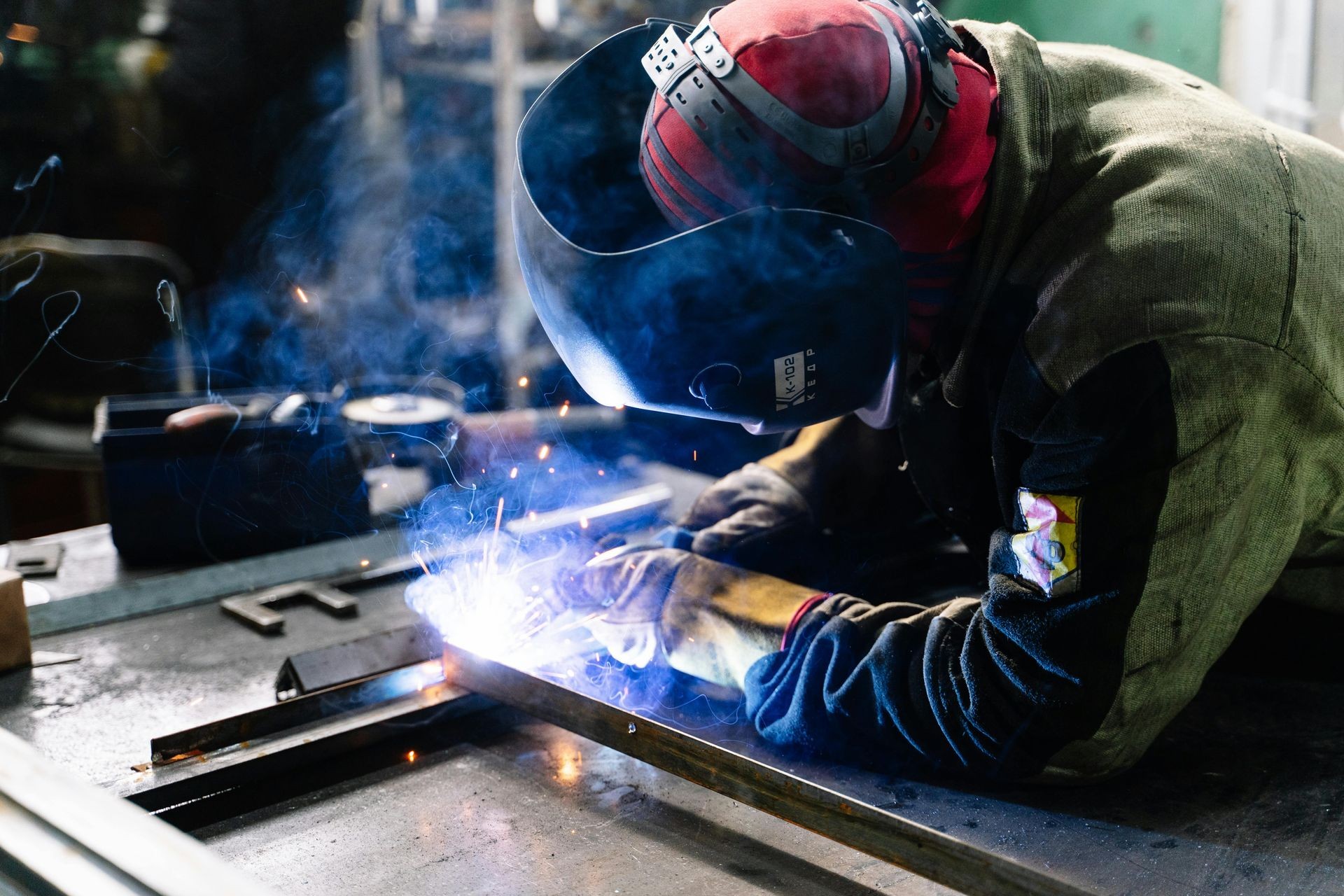 Welder wearing protective gear and helmet, working on metal with bright sparks and blue smoke.