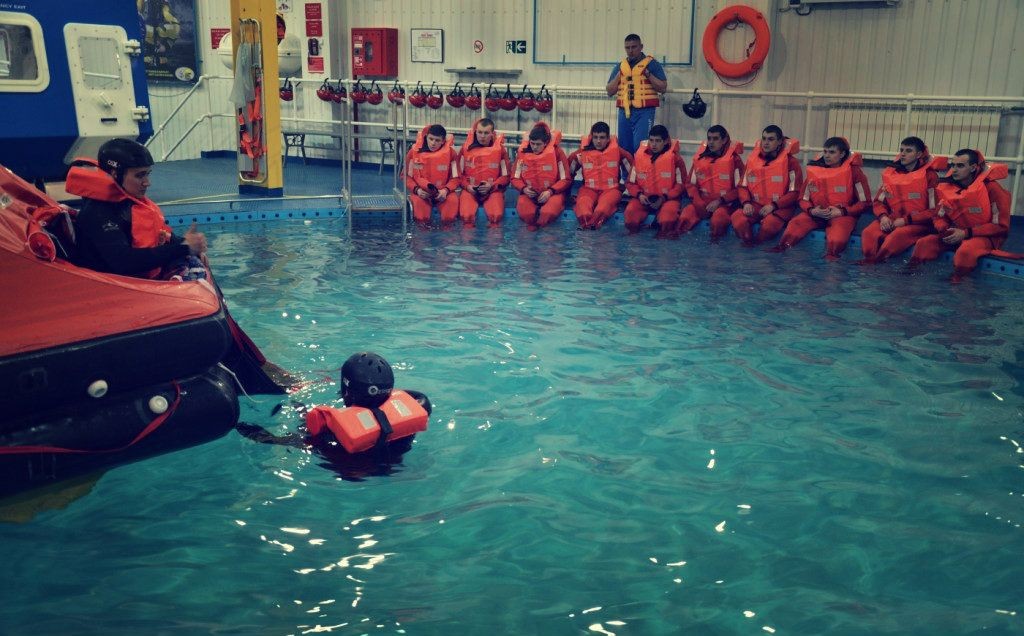 Group of people in orange life jackets sitting by a pool during water safety training session.