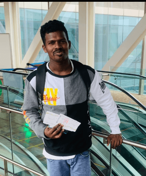 Man standing in an airport holding a passport and boarding pass, wearing a casual sweatshirt and backpack.