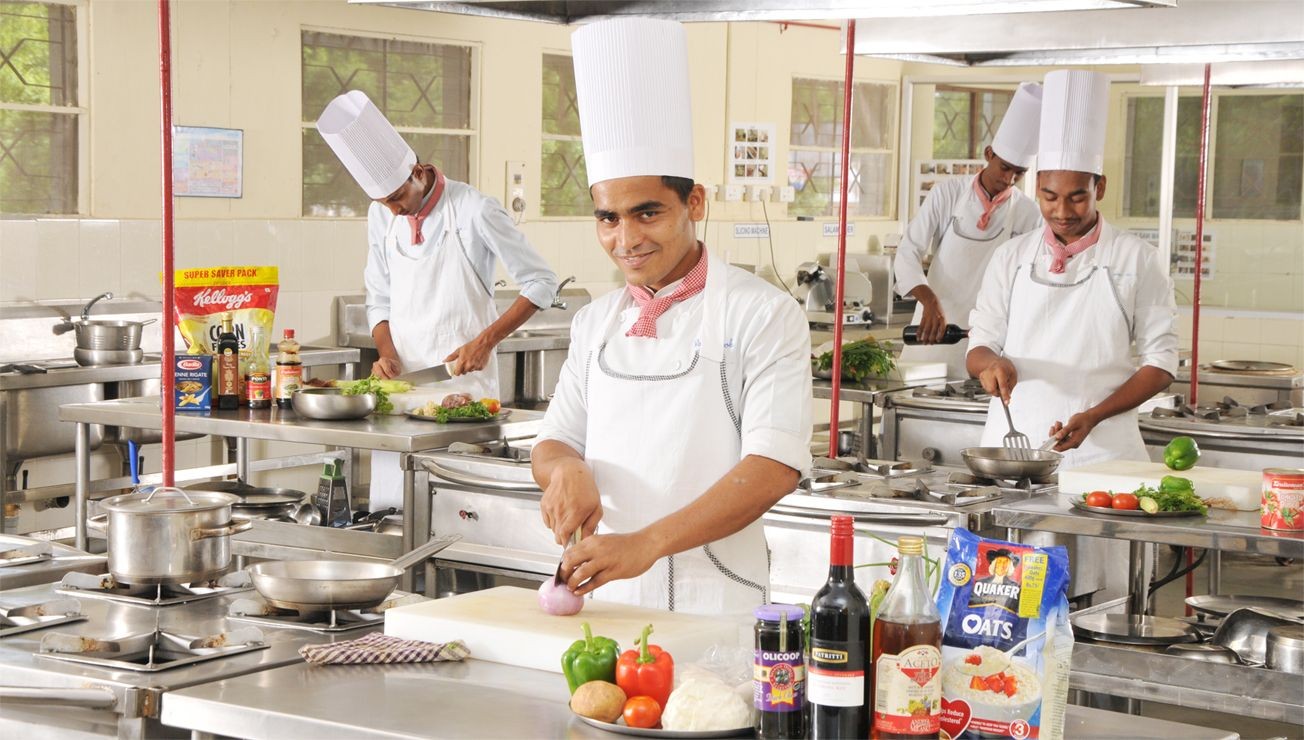 Chefs in a commercial kitchen preparing various dishes, with fresh vegetables and ingredients on the countertops.